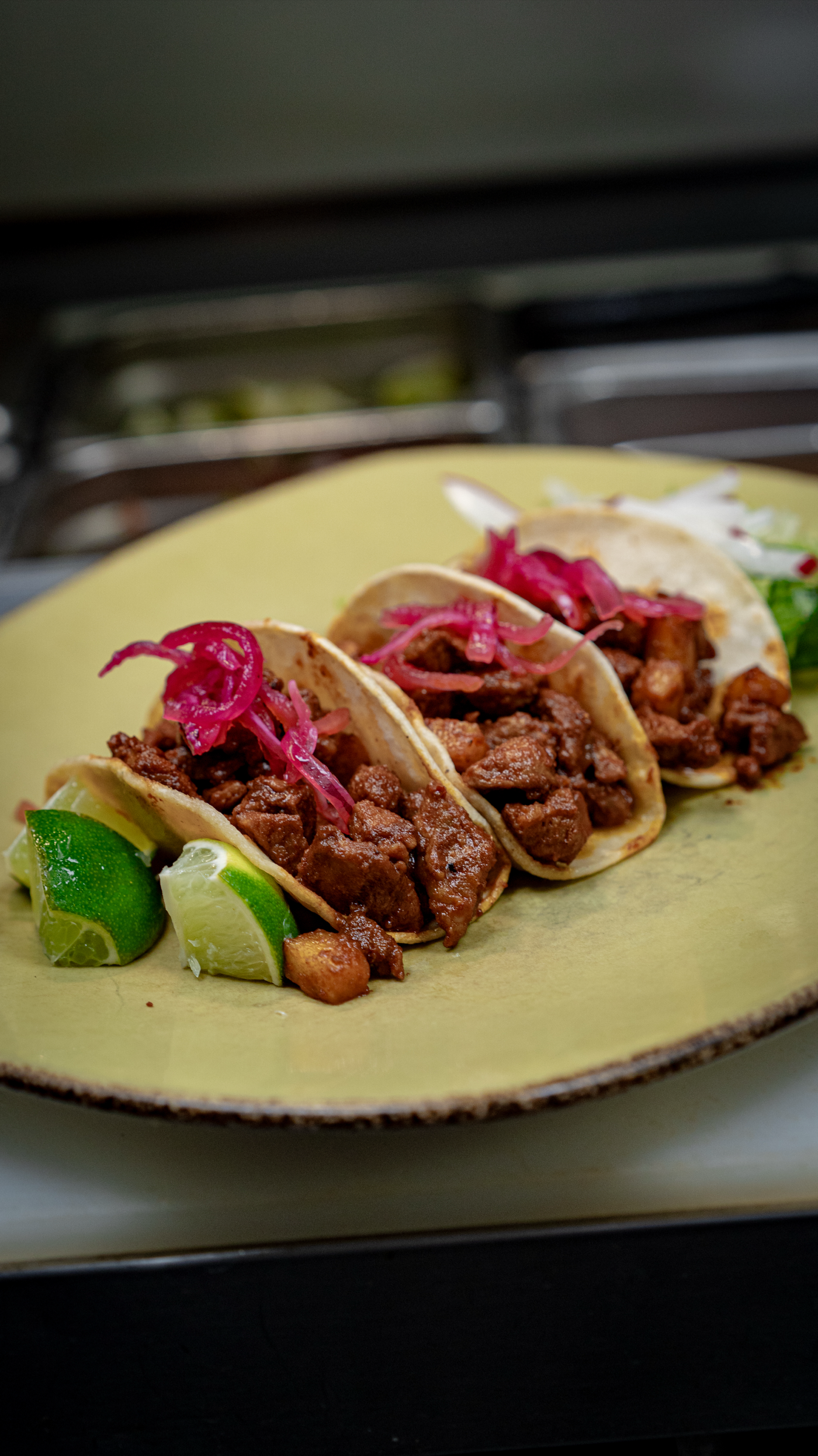 Three al pastor tacos on a rustic ochre ceramic plate with pickled red onion and lime.
